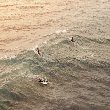 HUNTINGTON BEACH SURF: Three surfers are riding waves at sunset. The surfers are wearing wetsuits and are paddling on their surfboards. The waves are breaking in the distance and the sun is setting in the background, casting a golden glow over the water.