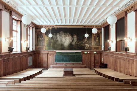 LA SORBONNE AMPHITHEATRE: A view of a large auditorium with wooden benches, a mural on the back wall, and a high ceiling with lights. The room is empty, suggesting it may be before or after a lecture or event.