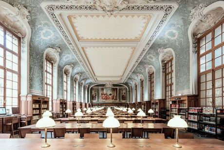 LA SORBONNE LIBRAIRIE: This image showcases the interior of a grand library reading room in France. The room is characterized by high ceilings adorned with intricate molding and a light blue and white color scheme. Large windows line the walls, allowing natural light to illuminate the rows of wooden desks and bookshelves that fill the space. Lamps on each desk provide additional light for readers.