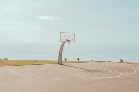 LA PARK: An empty basketball court with a hoop and net is situated on a grassy hill overlooking a vast ocean. The court is made of concrete and the sky is a clear blue, suggesting a sunny day. The image evokes a sense of tranquility and solitude, perfect for a game of one-on-one or simply enjoying the view.