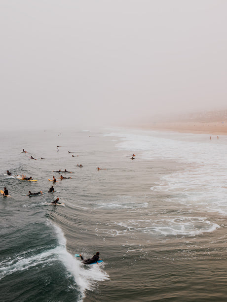 LONG BEACH SURF: An aerial view of surfers riding waves in foggy coastal waters. The surfers are spread out across the water, with some catching waves and others paddling. The fog creates a hazy and ethereal atmosphere.