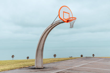 LOS ANGELES BASKETBALL: A basketball hoop stands tall on a paved court overlooking a blue ocean on a cloudy day. The hoop is in the foreground, and the ocean stretches out to the horizon. There are a few palm trees in the background, and the sky is a pale blue.