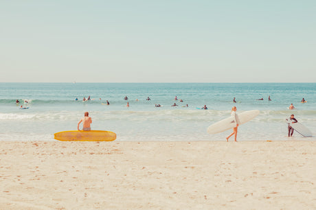 LOS ANGELES SURF: Two surfers are walking along a sandy beach with their surfboards after a day of surfing. The ocean is in the background with many other surfers in the water. The weather is sunny and the sky is blue.