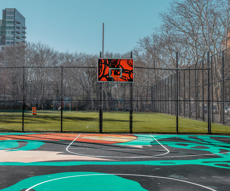 NEW YORK BASKETBALL COURT 2: A view of a basketball court on a sunny day, with a black chain-link fence surrounding the court. The court is made of artificial turf with bright green and orange painted lines. The backboard is a unique design with bright orange and black accents. There are trees and buildings in the background, suggesting an urban location.