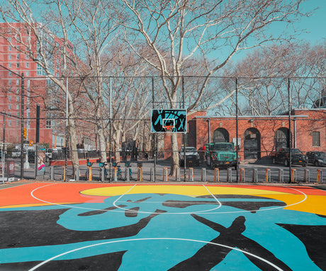 NEW YORK BASKETBALL COURT: A vibrant basketball court with a colorful design sits in a New York City park, surrounded by trees and a brick building. The court is enclosed by a chain link fence, and there are several parked cars in the background. The sun is shining brightly, and the sky is a clear blue.