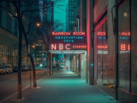 NEW YORK NBC: A vibrant neon sign advertising the Rainbow Room Observation Deck at NBC Studios illuminates an otherwise empty street in New York City at night. The sign glows brightly, casting a colorful light on the sidewalk and the surrounding buildings. The street is deserted, with only a few cars parked along the curb. The scene is a mix of bustling city life and quiet solitude, capturing the unique atmosphere of New York City after dark.