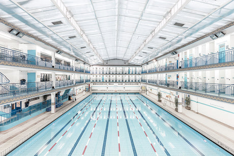 PISCINE PONTOISE: A photograph of an indoor swimming pool with multiple lanes. The pool is surrounded by white walls and has a balcony overlooking the water. The ceiling is high and has a skylight. There is a lot of natural light coming in through the windows.