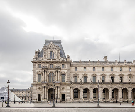 PAVILLON MOLLIEN LE LOUVRE: The iconic Louvre Museum in Paris, France, stands tall under a cloudy sky. The buildings intricate facade features ornate detailing and tall arched windows. The expansive courtyard in front of the museum is empty except for a few people walking by. The scene evokes a sense of historical grandeur and architectural beauty.