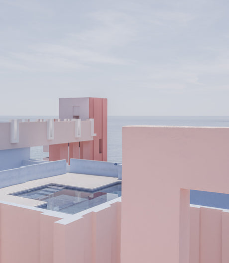 POOL WITH THE SEA: A modern building with a swimming pool on the roof is pictured against a bright blue ocean and a clear, pale blue sky. The building features white and pink walls and a flat roof design. The buildings structure is unique and geometric with clean lines and sharp angles.