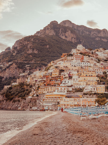 POSITANO SUNSET: A picturesque view of a coastal town in Italy, nestled against a backdrop of majestic mountains. The towns colorful buildings cascade down the hillside, reaching towards the sandy beach below. The setting sun casts a warm glow over the scene, highlighting the tranquility of the moment.