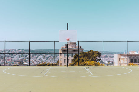 SFO BASKETBALL: This photo shows a basketball hoop standing on an outdoor court, with a chain-link fence surrounding it. The court is empty, but there is a beautiful view of a city skyline in the background, with buildings and trees visible. The sky is bright blue, suggesting a sunny day.