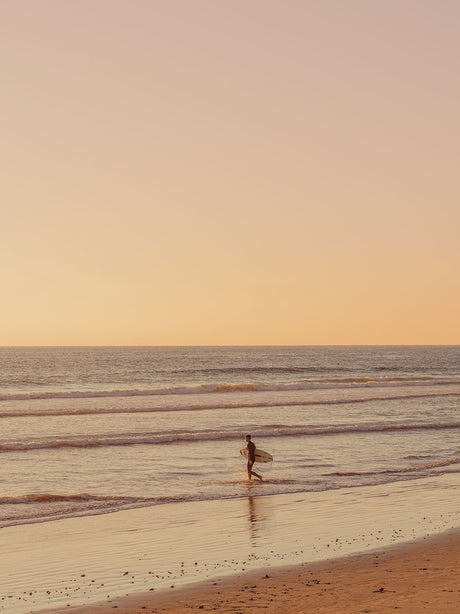 TAKE OFF: A lone surfer walks along the sandy beach, carrying a surfboard, as the sun sets over the ocean. The water is calm and the sky is a soft orange color. The surfer is silhouetted against the sunset.