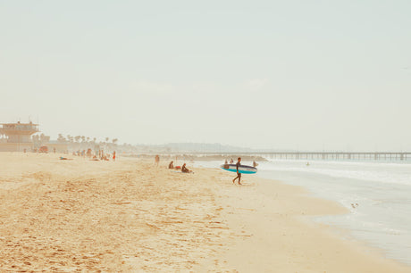 VENICE BEACH SURF: A surfer walks along a sandy beach towards the ocean. The surfer is carrying a surfboard. The ocean is calm with small waves. There are people swimming and sunbathing in the background.