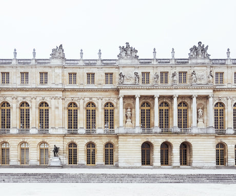 VERSAILLES 2: A wide shot of the Palace of Versailles in France, with its facade covered in snow. The ornate architecture, statues, and windows are all visible, and a single figure stands in the courtyard. The image captures the beauty of the palace in a winter setting.