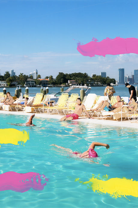 LA PISCINE: A scenic view of a resort pool in Miami Beach, Florida. The pool is surrounded by lounge chairs, with several people relaxing and swimming. The water is crystal clear and the sky is a vibrant blue. In the distance, there are buildings and boats on the water, making for a picturesque setting.