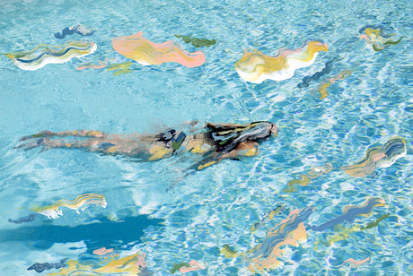 SWIM: A woman swims in a blue pool on a sunny day. The water is clear and the sun is shining brightly. The womans body is obscured by the water, but her hair is visible and she is swimming towards the camera. The water is rippling and reflecting the sunlight.