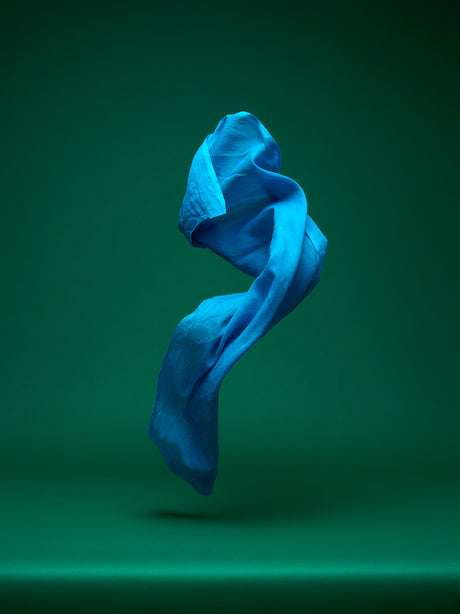 DANCING FABRIC, FLASH OF BLUE: A single, blue piece of fabric floats against a green background, the fabric is in a swirling and dynamic motion. The image is simple and elegant, highlighting the beauty of the fabrics movement.