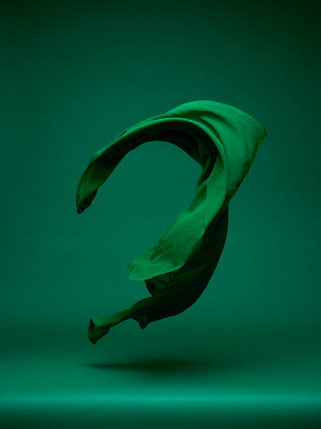 DANCING FABRIC, GREEN ON GREEN: A single piece of green fabric is shown floating through the air. The fabric has been twisted and turned to create an abstract shape. The fabric appears to be a soft, lightweight material that is easily moved by the air. The background of the image is a solid green color, which creates a sense of depth and contrast.