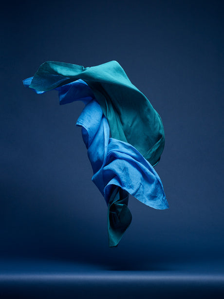 DANCING FABRIC, LIGHT BLUE AND GREEN: A photograph of blue and green fabric draped in mid-air against a dark blue background. The fabric is flowing and dynamic, creating a sense of movement and energy. The dark blue background provides a stark contrast to the bright colors of the fabric, highlighting its beauty and texture.