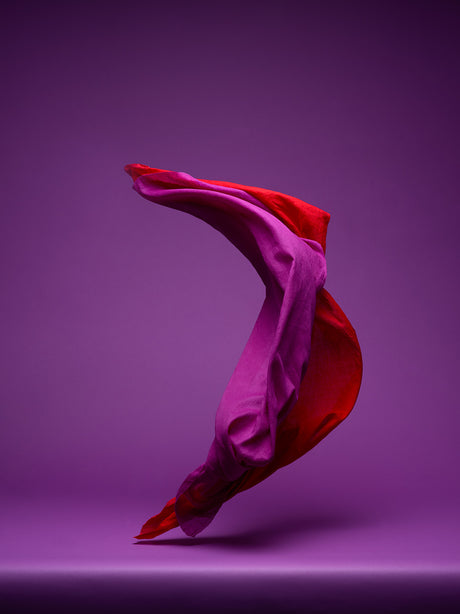 DANCING FABRIC, PURPLE AND RED: A close-up image of red and purple fabric draped in a dynamic, flowing motion against a solid purple backdrop. The fabric is textured and appears soft and delicate, creating a sense of movement and fluidity. The vibrant colors and simple composition create a visually striking and abstract image.