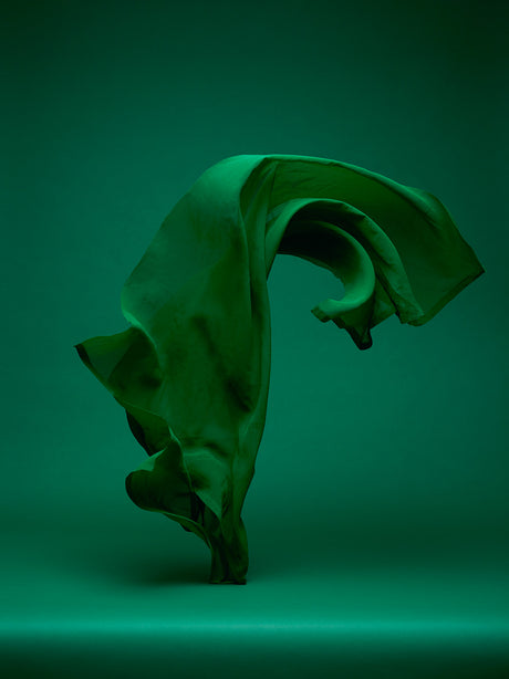 DANCING FABRIC, GREEN ON GREEN 2: A single piece of emerald green fabric swirls and floats in a dramatic fashion, creating a dynamic and abstract shape against a dark green backdrop. The image captures the movement and fluidity of the fabric, highlighting its texture and drape.