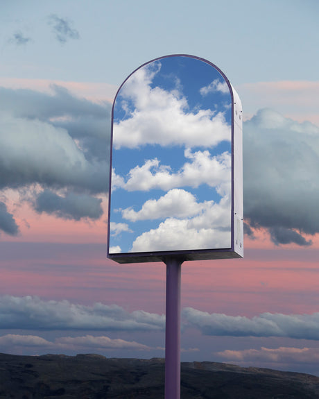 EARLIER: A single, tall, metal pole with a rectangular, arched mirror mounted on top reflects a clear blue sky with white fluffy clouds. The mirror is positioned in the foreground, facing the viewer. The background features a pastel pink sunset with dark clouds.