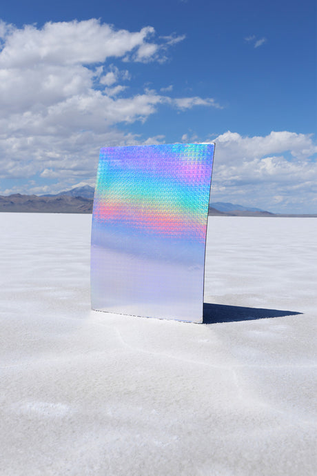 DID IT FIRST: A large, holographic panel stands upright on a white salt flat under a blue sky with white clouds. The panel is iridescent and reflects the colors of the sky and the surrounding landscape.