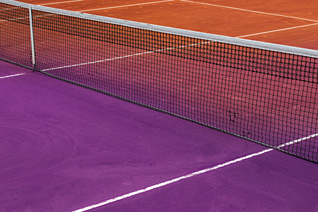 GAUDIO: A close-up image of a tennis court with a black net separating a purple side from a red clay side. The image is taken at an angle, highlighting the white lines that mark the courts boundaries.