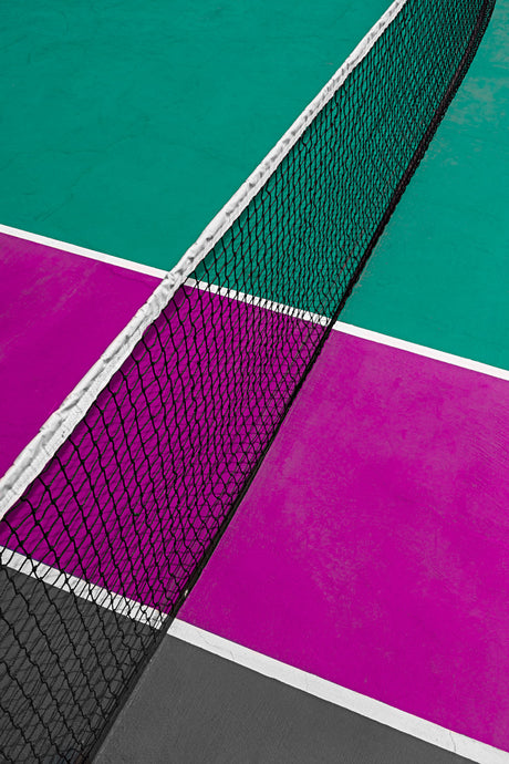 RAFTER: A close-up shot of a tennis net on a court with green, pink, and grey surfaces. The net is positioned diagonally across the court, with the focus on the black netting and white border. The courts lines are visible, dividing the colors into distinct sections.