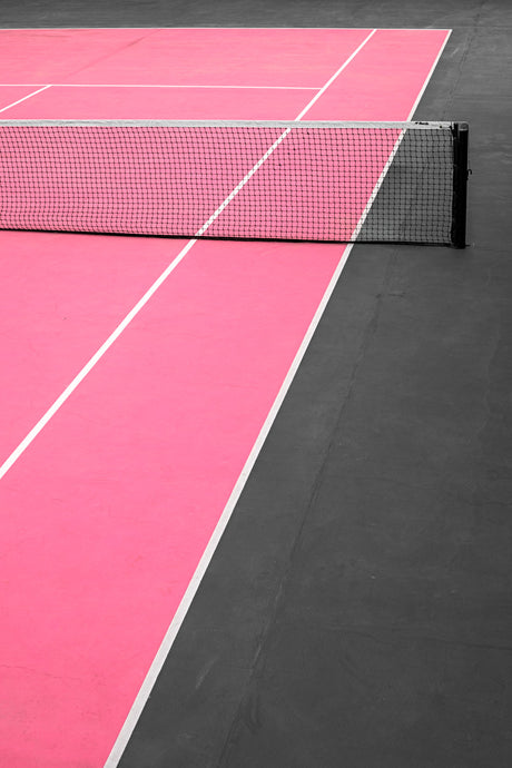 SELES: This image shows a pink tennis court with white lines and a black net. The net is partially visible, with the post extending to the right of the image. The court is brightly lit, with the pink surface reflecting the light.