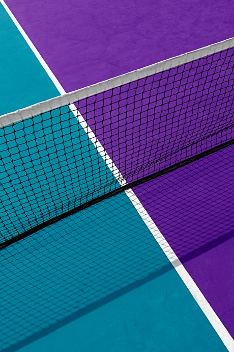 SVETLANA: A close-up shot of a black tennis net dividing a purple and a blue tennis court. The white lines of the court are visible on the sides. The net is in the foreground, with the court surfaces in the background.