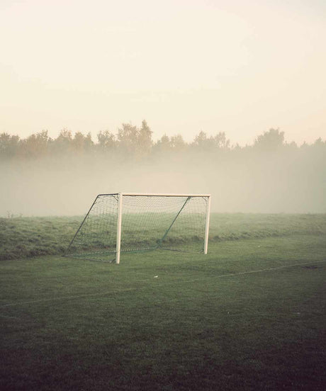 GOALS: A lone soccer goal stands on a grassy field, surrounded by a dense fog that obscures the trees in the background. The early morning light casts a soft, ethereal glow on the scene, creating a sense of tranquility and solitude.