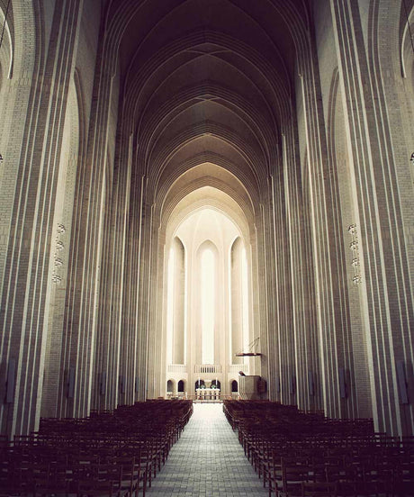 GRUNDTVIGS CHURCH: THE AISLE: An interior view of the Aarhus Cathedral in Denmark, showcasing the vastness and intricate details of the churchs architecture. The rows of pews lead towards the altar, bathed in the soft light streaming through the large windows. The high vaulted ceilings, constructed of brick and stone, create an air of grandeur and solemnity.