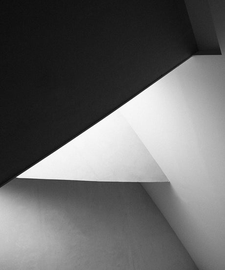 HEART 02: This is a black and white photograph that captures abstract geometric patterns formed by the architectural details of an interior space. Light and shadow play a key role in highlighting the simple lines and angles of the structure.