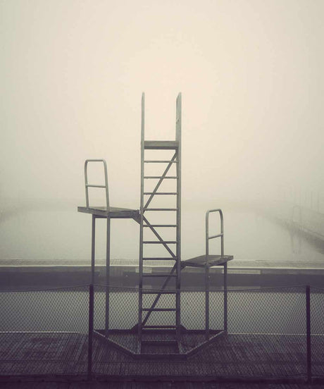 DESERTED CITY: POOL CHAIRS: A metal diving platform stands alone in the thick fog on a cold morning. The ladder leading up to the platform is visible in the center of the frame. The platform and ladder are surrounded by a chain-link fence.