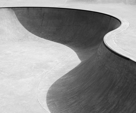 SKATE 01: A grayscale photo of an empty concrete skate park bowl. The smooth, curved surface of the bowl is illuminated by the afternoon sun, creating a stark contrast of light and shadow. The bowl appears ready for action, as skaters prepare to carve and grind their way through the park.