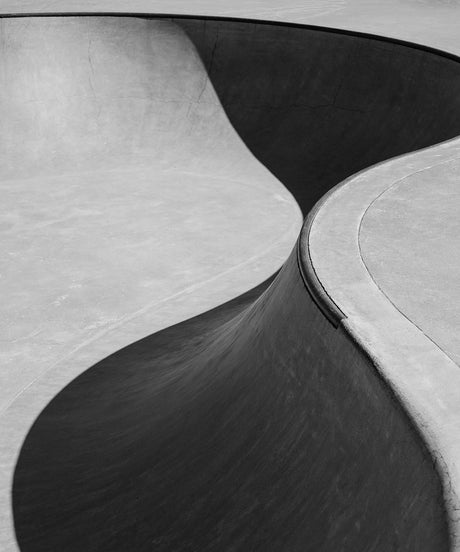 SKATE 02: A black and white image showcasing the detail of a concrete skate park ramp, captured in a close-up shot. The curved and textured surface of the ramp highlights the design and functionality of this outdoor recreation area.