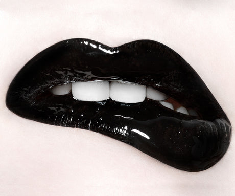 INK: This image is a close-up of a persons glossy black lips. The lips are slightly parted, revealing a glimpse of white teeth. The image is taken in a studio setting and has a shallow depth of field.