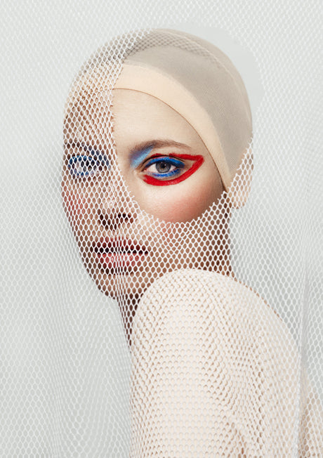 DECONSTRUCTED II: This image features a woman with blue and red eye makeup posing behind a white mesh fabric. Her gaze is directed slightly to the left, and her expression is composed. The image is likely a fashion editorial or beauty shoot, highlighting the bold eye makeup look.