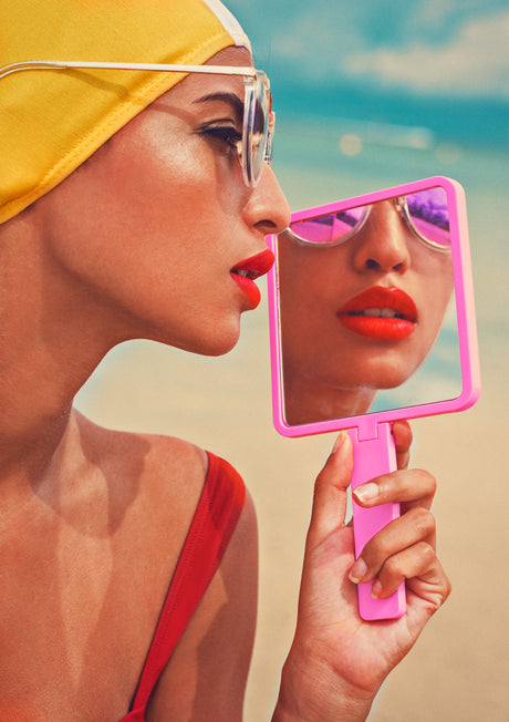 PINK MIRROR: This image depicts a young woman wearing a yellow swim cap and sunglasses, looking at herself in a pink hand mirror. She is standing on a beach, with the ocean behind her. The woman is wearing a red swimsuit and has a red lip.