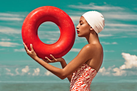 THE BLUE LAGOON AND THE RED CIRCLE: A woman wearing a white swim cap stands on a beach, holding a large, red inflatable ring in front of her. The woman is wearing a red and white swimsuit and has a tanned complexion. The background features a blue sky with white clouds and a turquoise ocean.