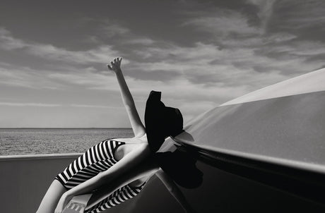 SUN BLOCK 1: A woman wearing a black and white striped swimsuit reclines on a boat deck, her hand reaching up toward a cloudy sky. The photo is in black and white, emphasizing the contrast between the woman, the boat, and the sky. The womans pose suggests relaxation and enjoyment of the open sea.