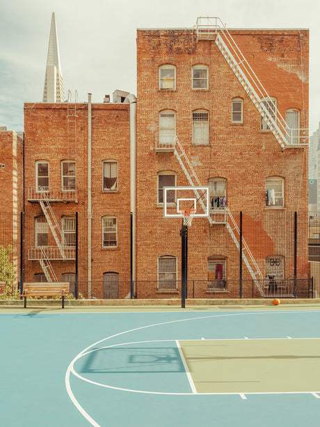 SAN FRANCISCO BASKETBALL COURT: A basketball court with a blue surface and white lines is situated in front of a red brick building. The building has fire escapes and windows, and there is a bench to the left of the court. The court is surrounded by a black fence, and there is a view of the city in the background. The photo captures a sunny day in San Francisco.