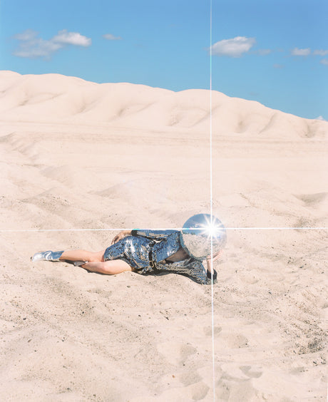 DISCO GIRL 1: A woman wearing a silver sequined dress lies on a sandy dune with a disco ball on her head. The sun is shining brightly and the womans head is reflecting light back at the camera.