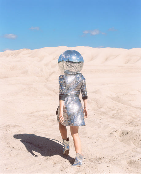 DISCO GIRL 2: A woman walks through a desert landscape wearing a shiny, silver dress and a disco ball helmet. The bright, blue sky and white sand dunes create a stark contrast with the womans outfit, making her appear like a futuristic vision in the middle of a desolate landscape.