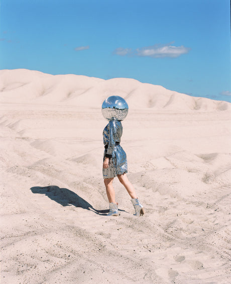 DISCO GIRL 3: A woman wearing a silver sequined dress walks across a sandy desert. She has a disco ball on her head and her shadow stretches out behind her. The sky is clear blue and the desert is empty except for the woman.