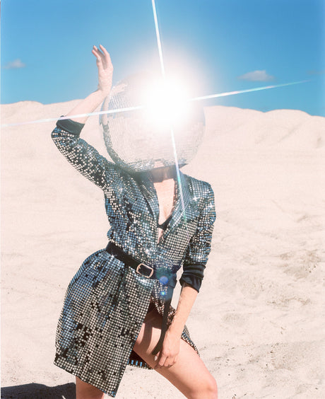 DISCO GIRL 4: A woman wearing a sequined jacket and a disco ball on her head stands in a desert landscape. The sun is shining brightly, creating a glare that obscures her face. She is wearing a black belt around her waist, and the light reflects off of the sequins in her jacket.