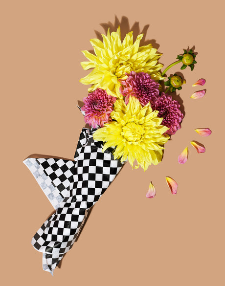 DELI DAHLIAS: A bouquet of yellow and pink dahlias are arranged in a checkered paper wrapping on a tan background. The bouquet is lying on its side, and some of the petals have fallen out of the wrapping. The image has a simple, clean aesthetic.