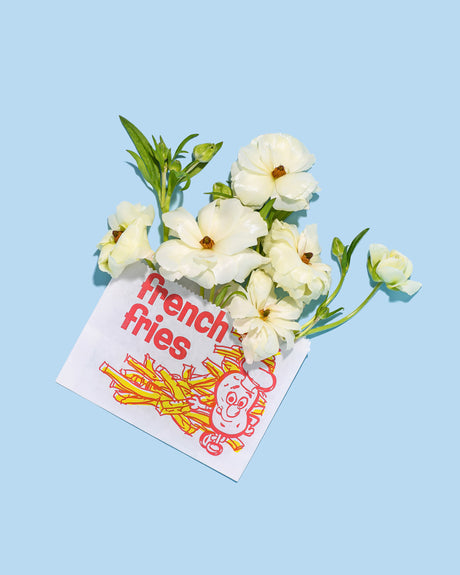 FRENCH FRY RANUNCULUS: This image shows white flowers with green stems arranged in a french fries paper bag. The paper bag has red and yellow text and a cartoon image of a potato with a chefs hat. The background is a light blue.