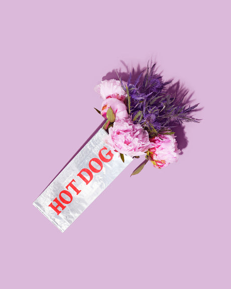 HOT DOG PEONIES: A bouquet of pink peonies and purple flowers is arranged in a hot dog paper wrapper, laying on a lavender-colored background. The flowers are soft and delicate, with the peonies in full bloom. The hot dog wrapper is white and has red lettering that says HOT DOG.